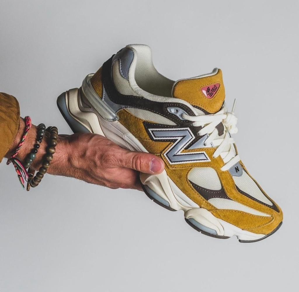 fcb40be3-2677-4967-b4b6-074ddb3accc5 | UAE SPORT STORE Person's hand with beaded bracelets holding a mustard-yellow and white New Balance sneaker against a plain gray background, side view.