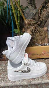 Pair of white Nike Air Force 1 sneakers with a silver decorative clip, resting on a granite surface beside a potted plant.