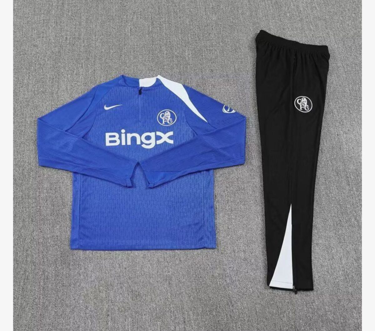 Chelsea FC blue training jersey with BingX sponsor and white accents, paired with black club-crest track pants on a gray surface