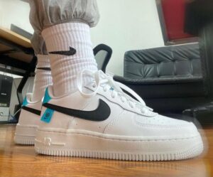 White Nike Air Force 1 sneakers with a black swoosh, teal heel tab, and blue tag, worn with white ribbed socks on a wooden floor.]}