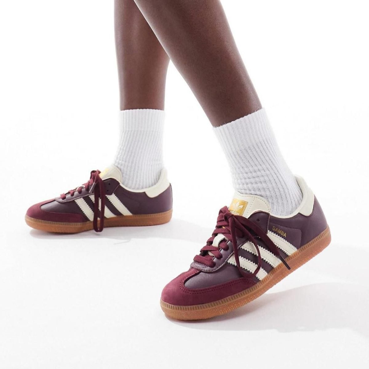 Maroon Adidas Samba sneakers with white stripes and gum sole, worn with white socks against a white background.