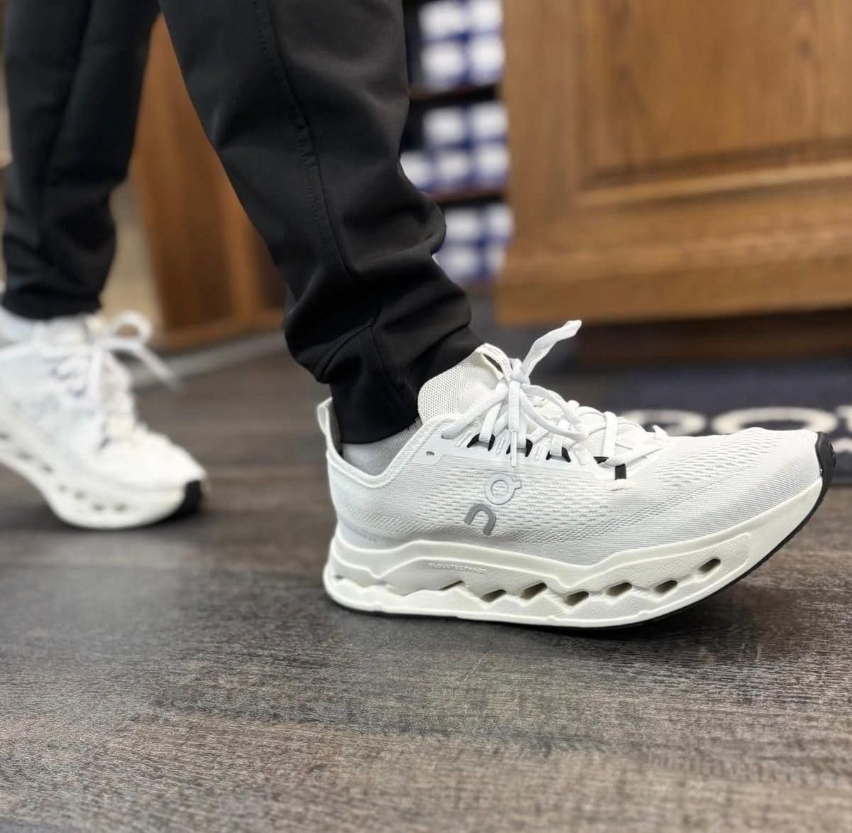 Close-up of a white athletic sneaker with a chunky sole on a foot in black pants, standing on a dark floor.