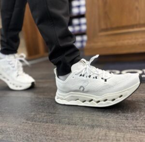 Close-up of a white athletic sneaker with a chunky sole on a foot in black pants, standing on a dark floor.