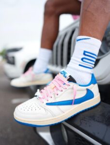 Close-up of a white Nike Air sneaker with pink laces and a blue swoosh, worn with white socks showing blue lettering/stripes on a raised surface.