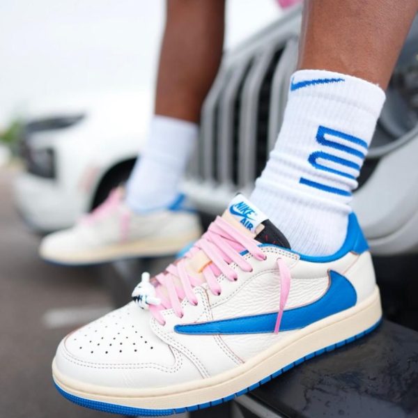 Close-up of a white Nike Air sneaker with pink laces and a blue swoosh, worn with white socks showing blue lettering/stripes on a raised surface.