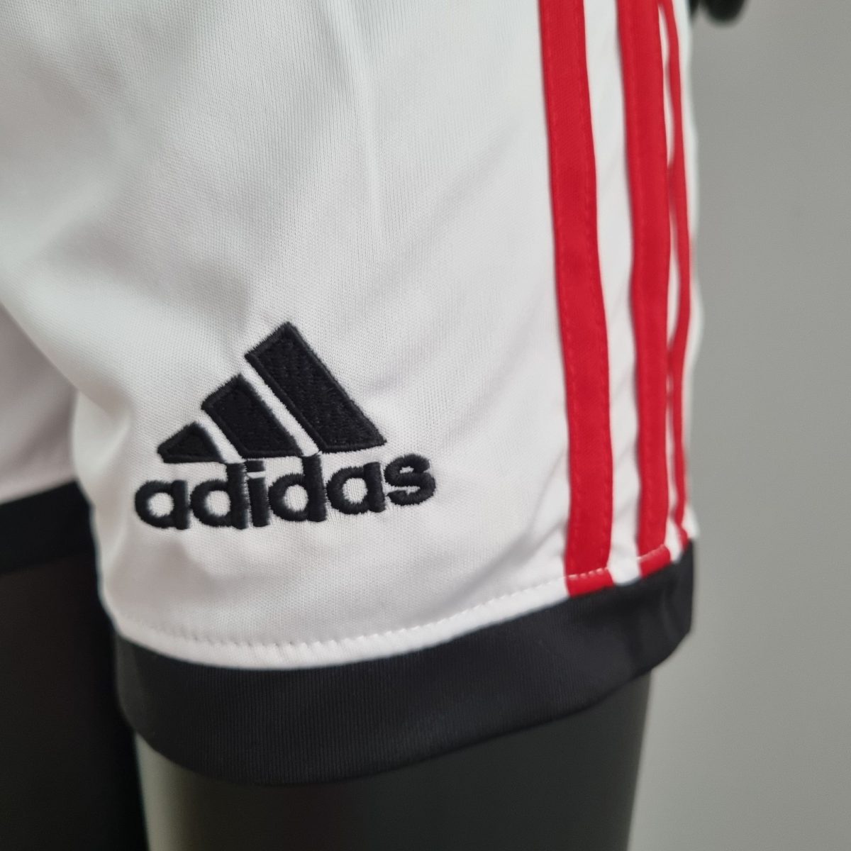 Close-up of white Adidas fabric showing the black Adidas logo and red vertical stripes on the side of a garment, with a dark hem at the bottom.