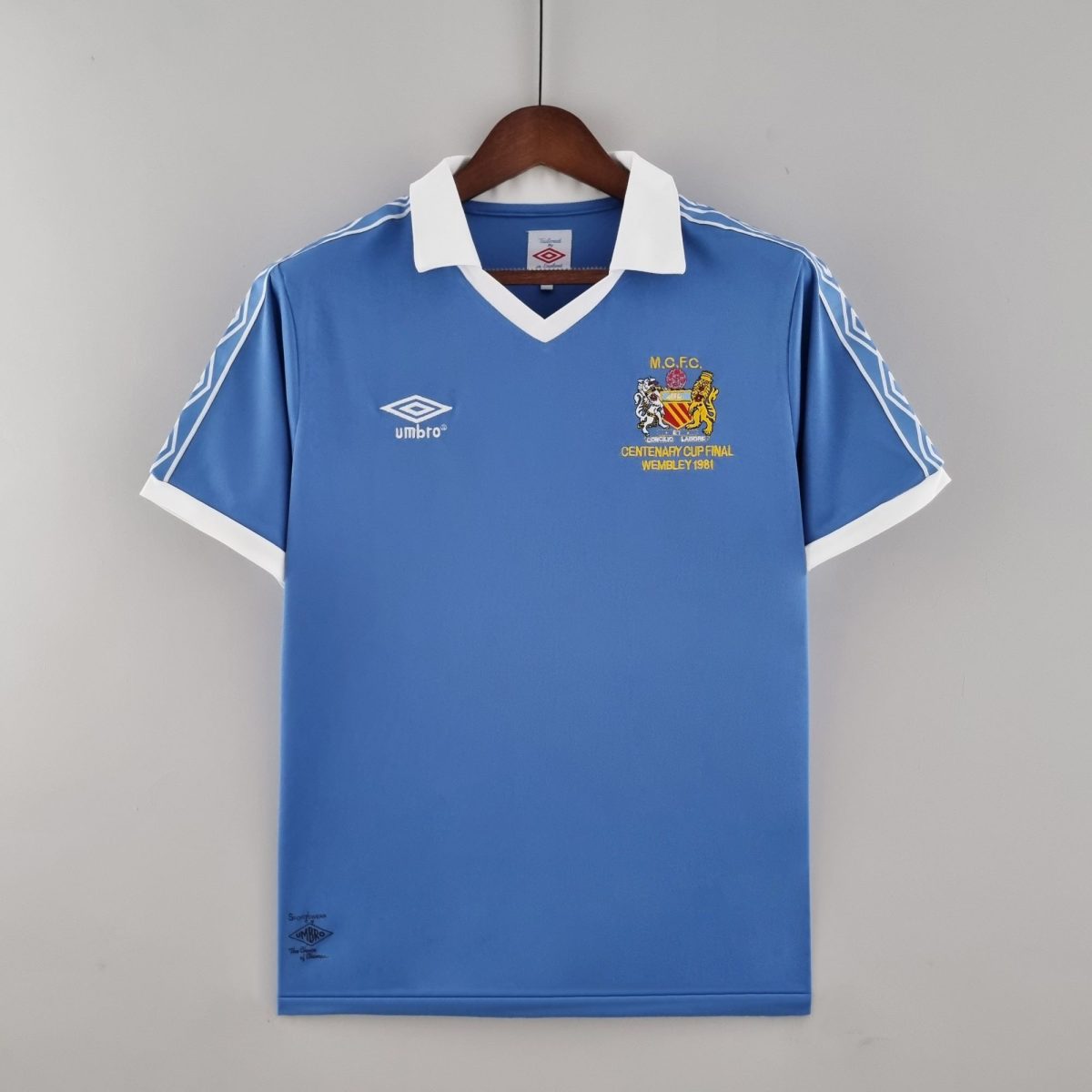 Blue Manchester City football shirt on a wooden hanger, white collar and cuffs, with Umbro branding and a crest reading 'MCFC Centenary Cup Final Wembley 1981' on the chest.