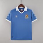 fd6e29d6_0338d15f-5377-4fa2-9bb4-a77f8238d85b | UAE SPORT STORE Blue Manchester City football shirt on a wooden hanger, white collar and cuffs, with Umbro branding and a crest reading 'MCFC Centenary Cup Final Wembley 1981' on the chest.