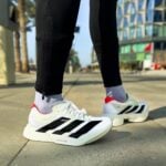 Close-up of white Adidas running shoes with black stripes on a sidewalk, wearing gray socks and black leggings.