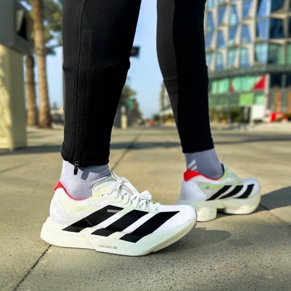 Close-up of white Adidas running shoes with black stripes on a sidewalk, wearing gray socks and black leggings.