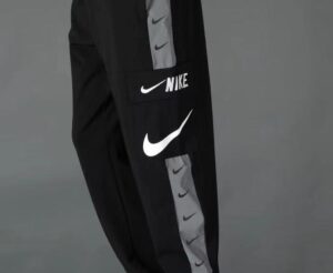 Close-up of black Nike track pants with a gray side stripe and white swoosh logos along the leg, including a pocket with 'NIKE' text.