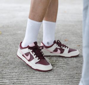 fdb7e2ab-d971-4990-9d96-55da49987866_1 | UAE SPORT STORE Burgundy and white sneakers with pink accents on a person wearing white crew socks, standing on a textured sidewalk surface.
