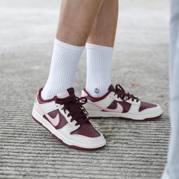 Burgundy and white sneakers with pink accents on a person wearing white crew socks, standing on a textured sidewalk surface.
