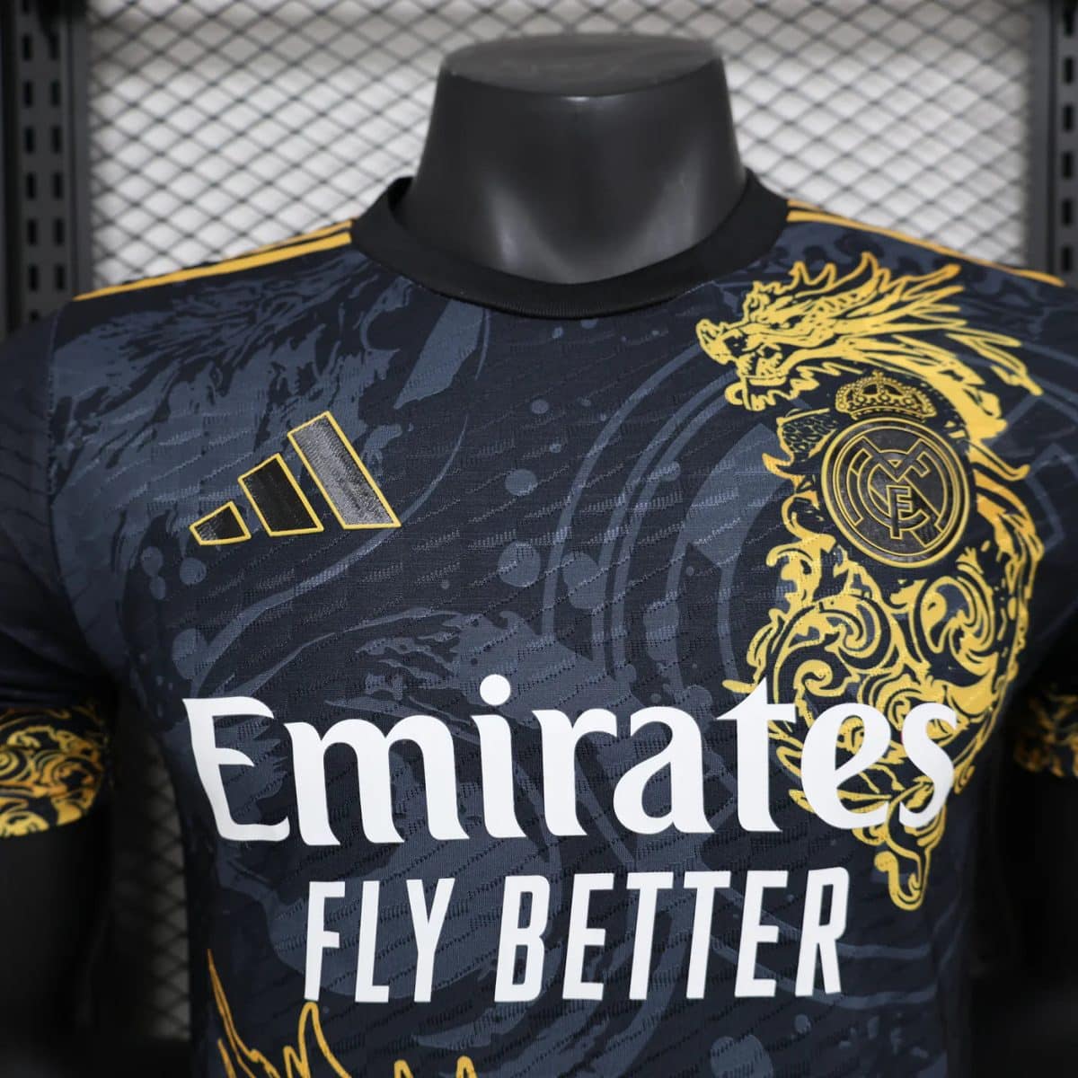 Close-up of a Real Madrid soccer jersey with a black‑gold pattern, crest on the right chest, and Emirates Fly Better slogan visible.