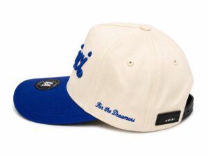 Beige snapback hat with a royal blue curved brim, blue 'For the Dreamers' embroidery, and a black AMIRI patch at the back.