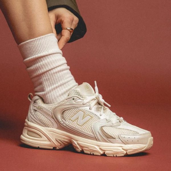 White New Balance sneaker with a chunky sole, worn with a white ribbed sock, against a terracotta background.