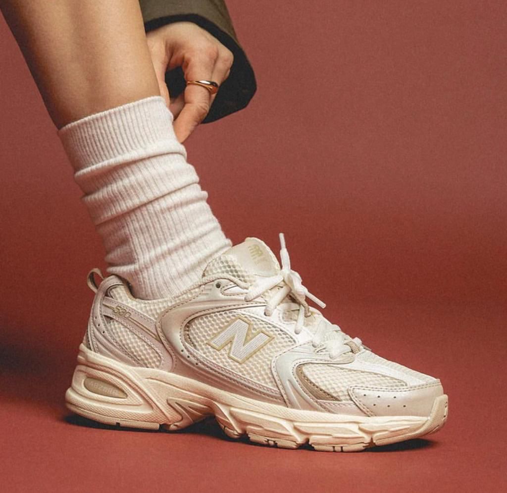 fdec7fb7-9e4c-431b-b196-4a02f8ff01e1 | UAE SPORT STORE White New Balance sneaker with a chunky sole, worn with a white ribbed sock, against a terracotta background.