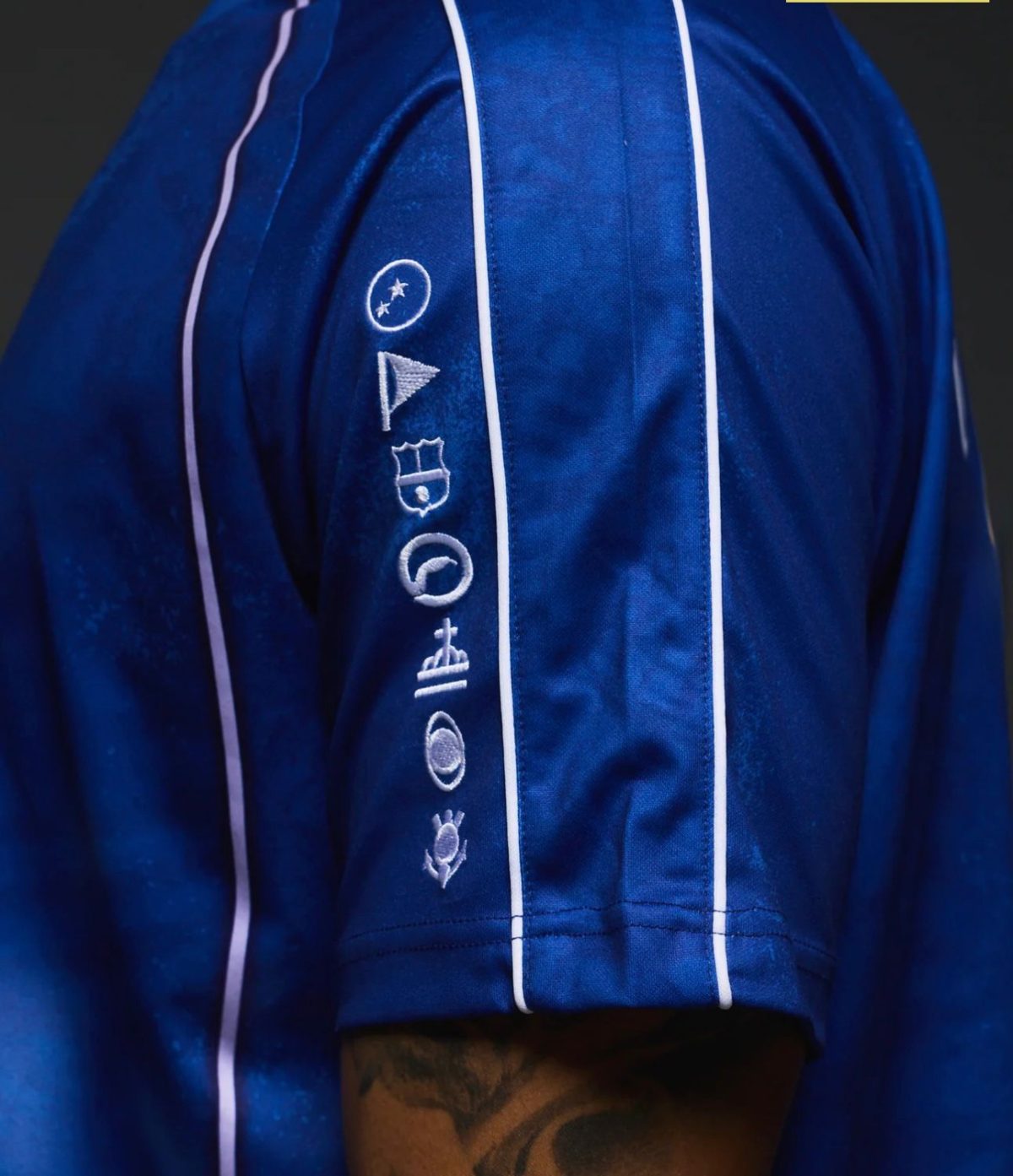 Close-up of a royal blue satin jacket sleeve with white piping and vertical embroidery of icons (circle, flag, shield, planet) along the forearm, with a tattooed arm visible at the bottom edge.