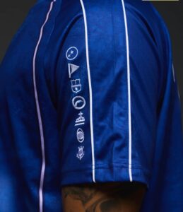 fe1eca33-818a-4ee7-b902-0765840b801e | UAE SPORT STORE Close-up of a royal blue satin jacket sleeve with white piping and vertical embroidery of icons (circle, flag, shield, planet) along the forearm, with a tattooed arm visible at the bottom edge.