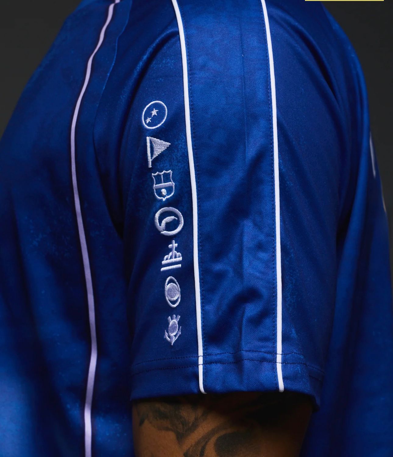 fe1eca33-818a-4ee7-b902-0765840b801e | UAE SPORT STORE Close-up of a royal blue satin jacket sleeve with white piping and vertical embroidery of icons (circle, flag, shield, planet) along the forearm, with a tattooed arm visible at the bottom edge.