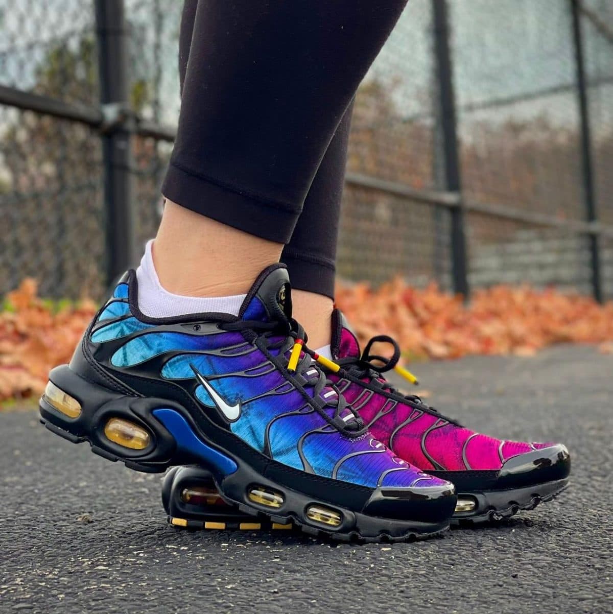 Close-up of colorful gradient running shoes (blue to pink) with a white Nike swoosh, standing on a paved path.