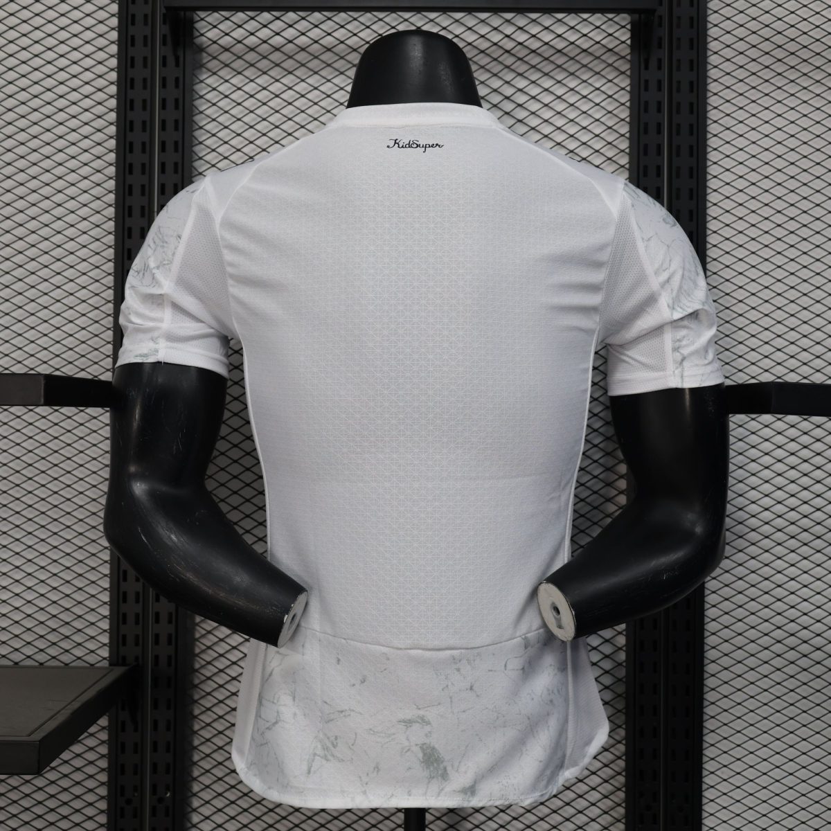 Back view of a white lattice-pattern sports jersey on a mannequin, with 'KidSuper' logo at the neck and a metal grid backdrop.