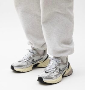 ffbe6d2c-4f03-47e3-a320-03356766046e | UAE SPORT STORE Close-up of feet in gray sweatpants wearing gray-white running shoes with a black Nike swoosh and cream soles.