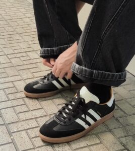 Person tying the laces of black sneakers with white stripes on a tiled sidewalk.