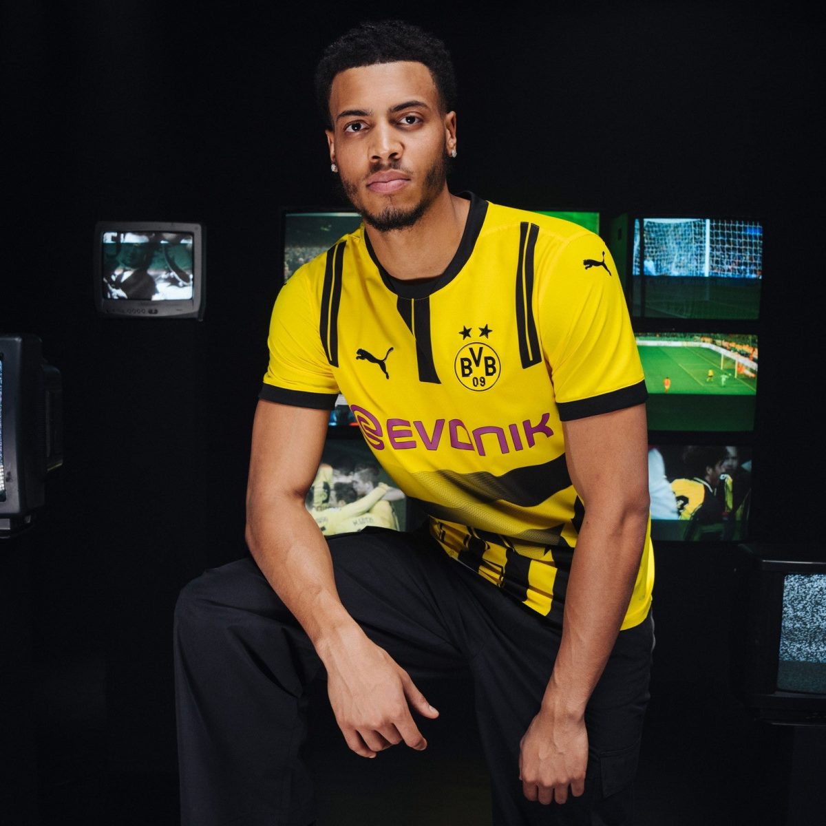 Portrait of a young man in a yellow Borussia Dortmund jersey posing in a dark studio with screens in the background.