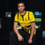 Portrait of a young man in a yellow Borussia Dortmund jersey posing in a dark studio with screens in the background.