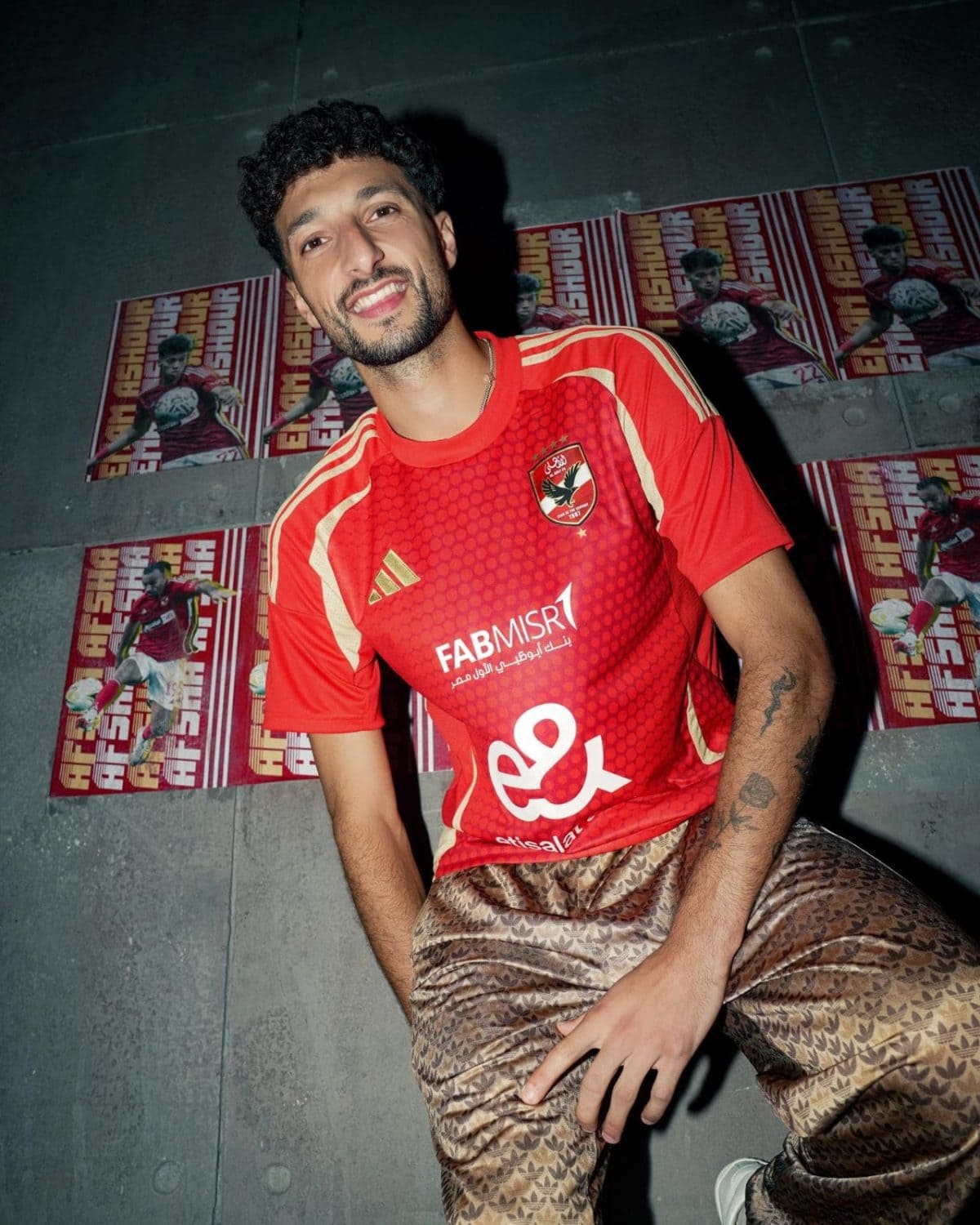 Young man in a red sports jersey leaning against a wall covered with posters, smiling at the camera.