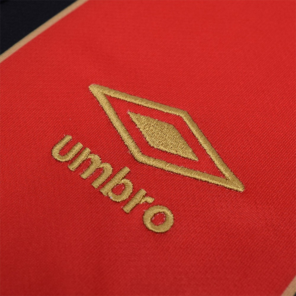 g7kJQvN0i0hobWT | UAE SPORT STORE Close-up of red fabric with gold embroidery of the Umbro logo and brand name.
