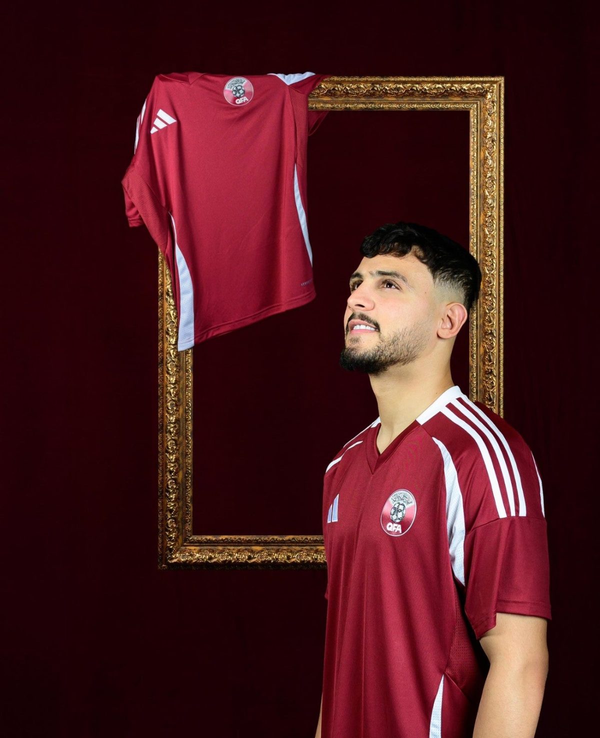 Man in a maroon sports jersey stands beside a gold ornate frame with a matching jersey draped over its top, against a dark background.
