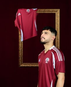 Man in a maroon sports jersey stands beside a gold ornate frame with a matching jersey draped over its top, against a dark background.