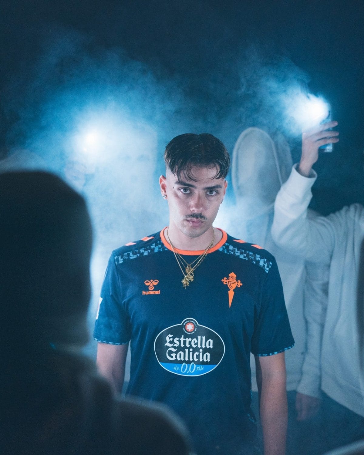 Young man in a blue Estrella Galicia jersey with gold chain stands in a smoky, blue-lit scene, looking at the camera.