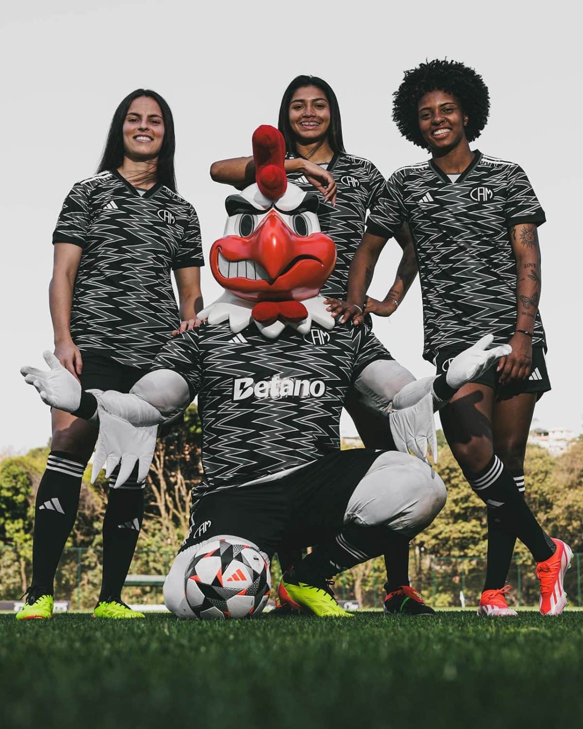 Three female soccer players pose with a large red chicken mascot on a grassy field, wearing matching black-and-white zigzag jerseys.