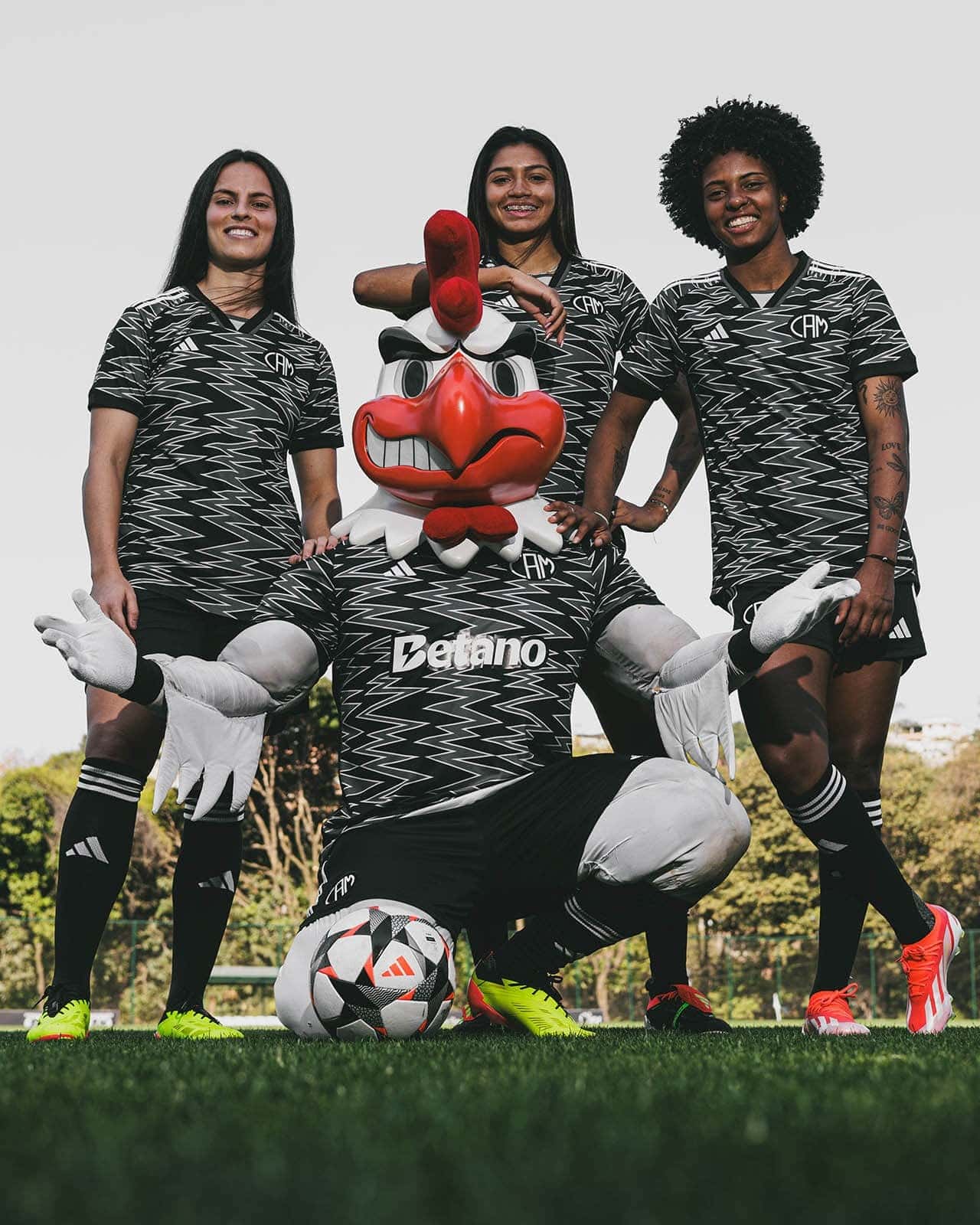 gCklYJHk6zgJTqo | UAE SPORT STORE Three female soccer players pose with a large red chicken mascot on a grassy field, wearing matching black-and-white zigzag jerseys.