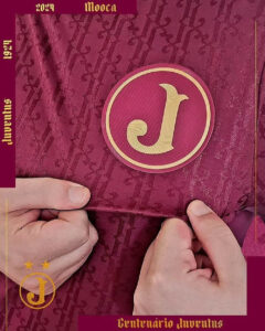 Close-up of a maroon cover with a gold circular J emblem; two hands are holding the edges as the cover is opened, with small gold text along the edges saying 2024 Moeda and Centenario Juventudes.