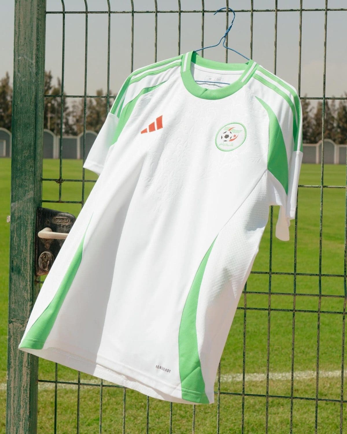 White and green soccer jersey hanging on a blue hanger from a metal fence, with a grassy field in the background and a team crest on the chest.