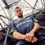 gPdfMEkauNic5S4 | UAE SPORT STORE Man in a dark blue MediaMarkt jersey stands under a metal stadium framework, arms crossed, looking at the camera.