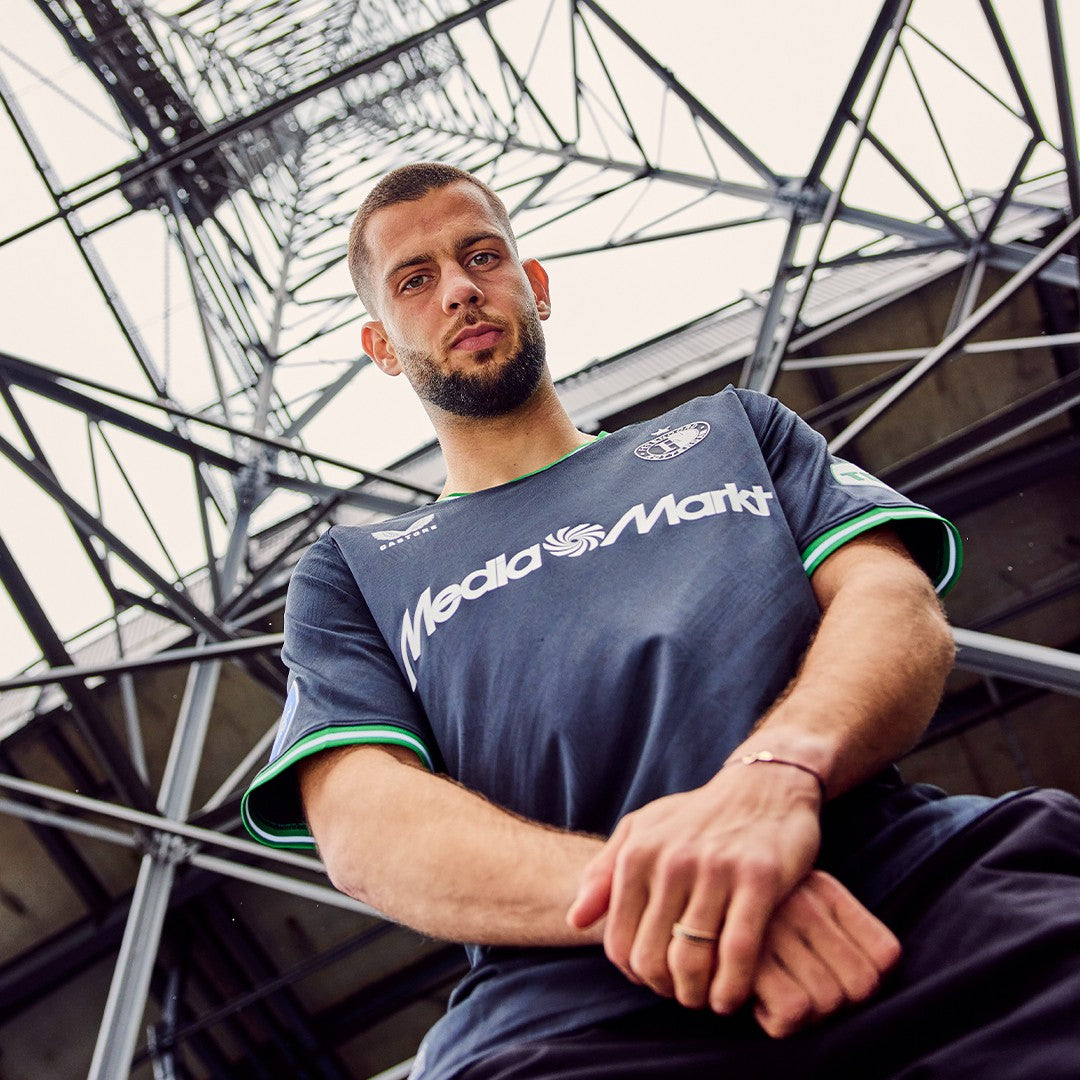 gPdfMEkauNic5S4 | UAE SPORT STORE Man in a dark blue MediaMarkt jersey stands under a metal stadium framework, arms crossed, looking at the camera.