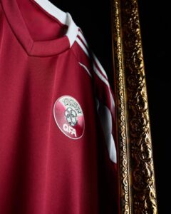 Close-up of a maroon sports jersey with white trim and the circular QFA emblem on the chest, beside a gold ornate frame.