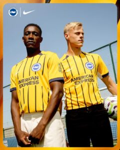 Two football players in yellow striped jerseys stand side by side outdoors, posing with a clear blue sky behind them and a green fence in the background.