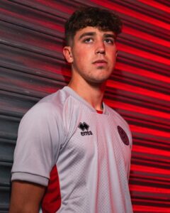 Portrait of a young man in a white team jersey with red side panels, standing against a red-lit striped background.