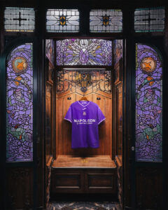 Purple Napoli soccer jersey hanging inside a vintage ornate elevator with intricate stained-glass panels surrounding it.