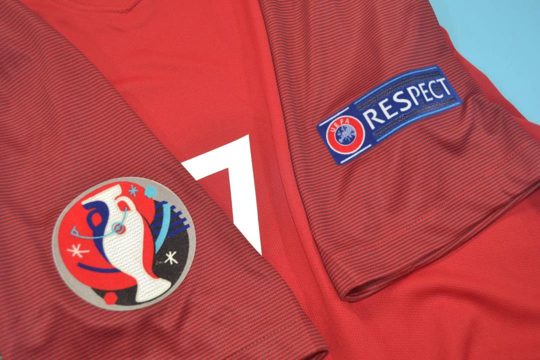 ghgfh_6ae82df8-057e-40aa-b442-801657cd86a3 | UAE SPORT STORE Red athletic jersey with a circular trophy emblem patch on one sleeve and a blue UEFA RESPECT patch on the other sleeve.