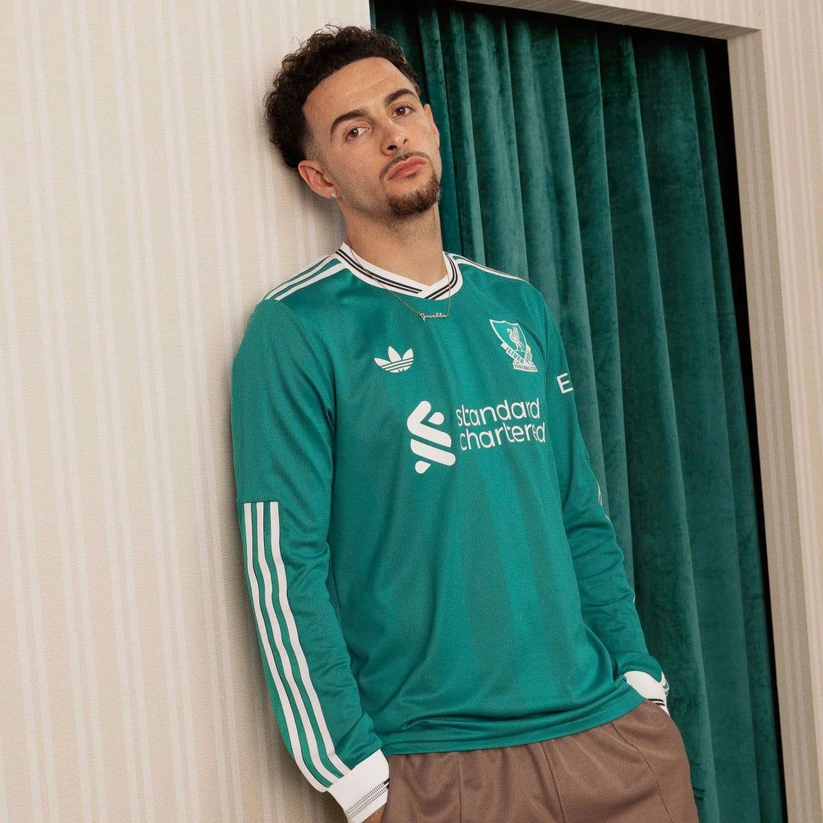 Young man leaning against a beige striped wall in a teal football jersey with sponsor logos visible on the front.