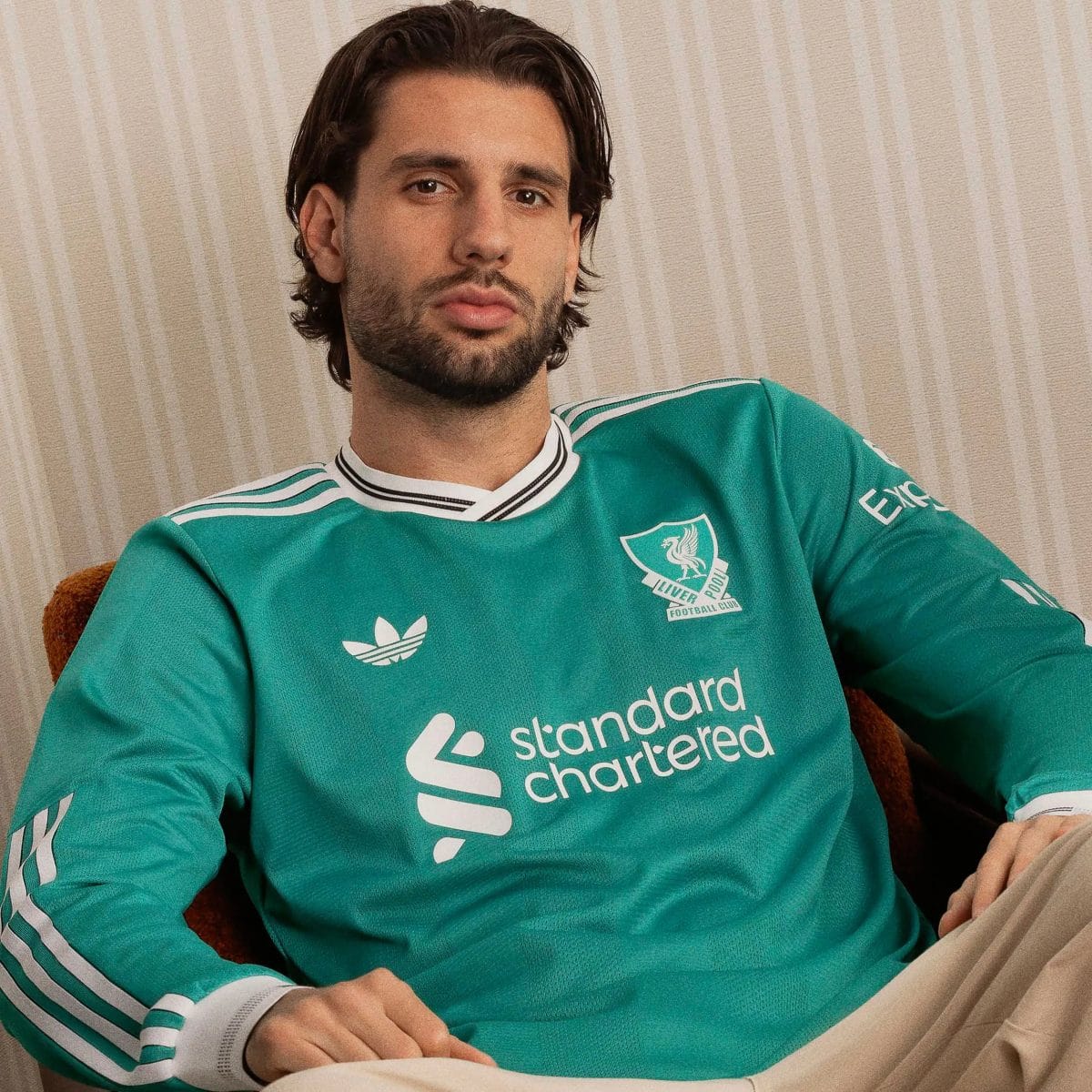 Man with dark hair and beard wearing a teal Liverpool FC jersey sits on a beige striped sofa, facing the camera.