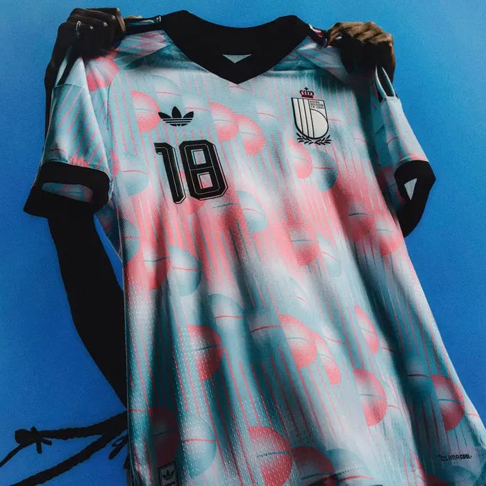 h24765-wc26_away_global_product_beauty_rbfa_2d_3x4_rgb-1385618 | UAE SPORT STORE Light blue-pink patterned soccer jersey with number 18 and crest, held up against a blue background by gloved hands.