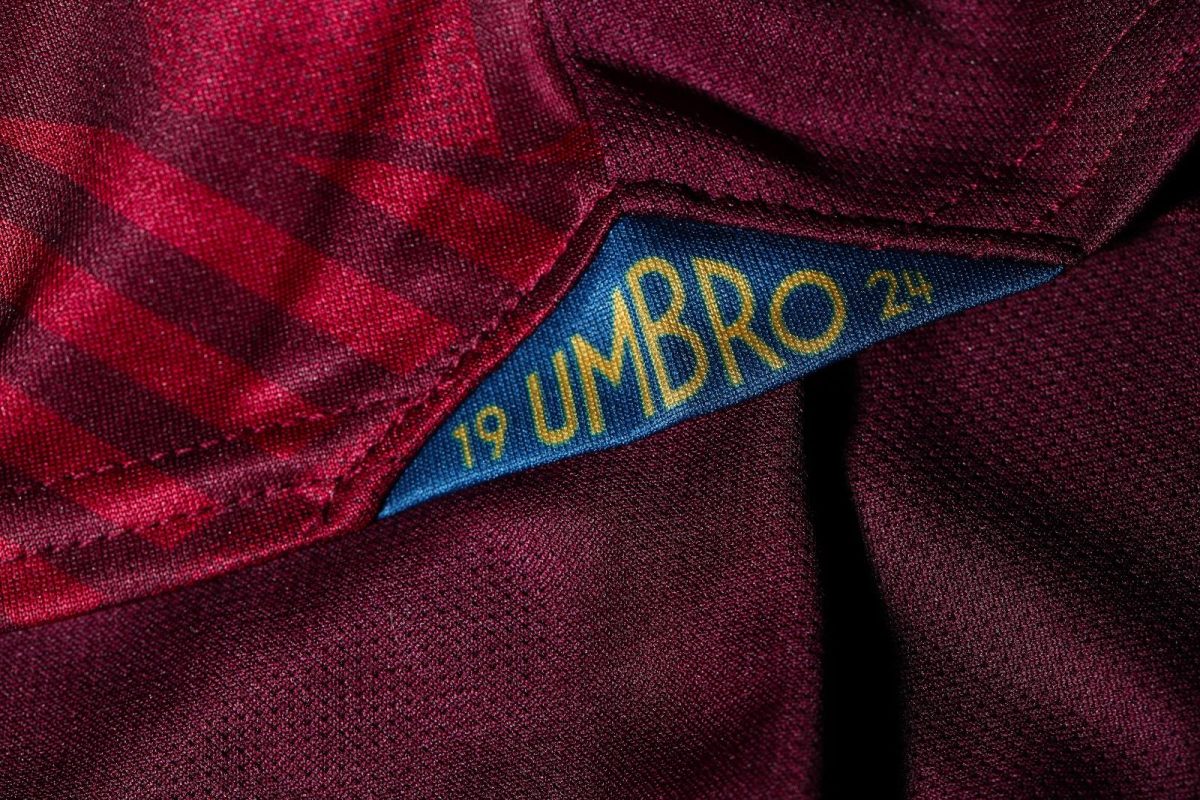 Close-up of a blue tag with yellow lettering reading 'UMBRO' tucked into a burgundy plaid fabric.